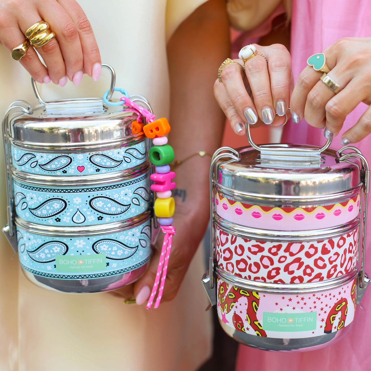 Boho-Tiffin stainless steel stackable lunchboxes with playful kiss, snake, and paisley prints held by two people