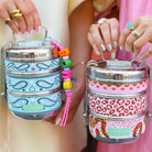 Boho-Tiffin stainless steel stackable lunchboxes with playful kiss, snake, and paisley prints held by two people